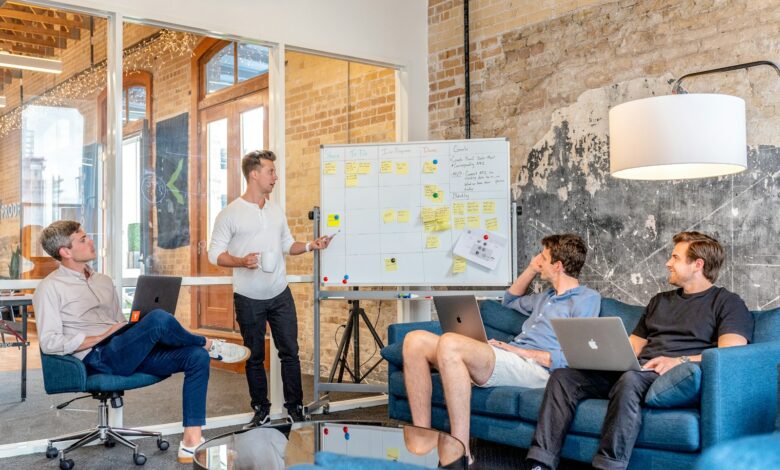 three men sitting while using laptops and watching man beside whiteboard